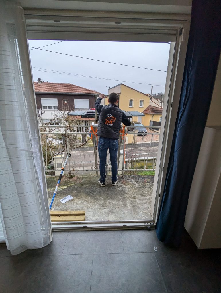 Un monte-meubles en cours d’utilisation devant une maison à étage. L’équipement, positionné sur l’allée pavée, permet de faire monter ou descendre des objets volumineux depuis un balcon situé au premier étage.