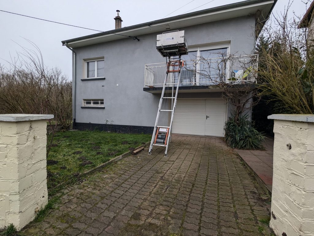 Un monte-meubles en cours d’utilisation devant une maison à étage. L’équipement, positionné sur l’allée pavée, permet de faire monter ou descendre des objets volumineux depuis un balcon situé au premier étage.